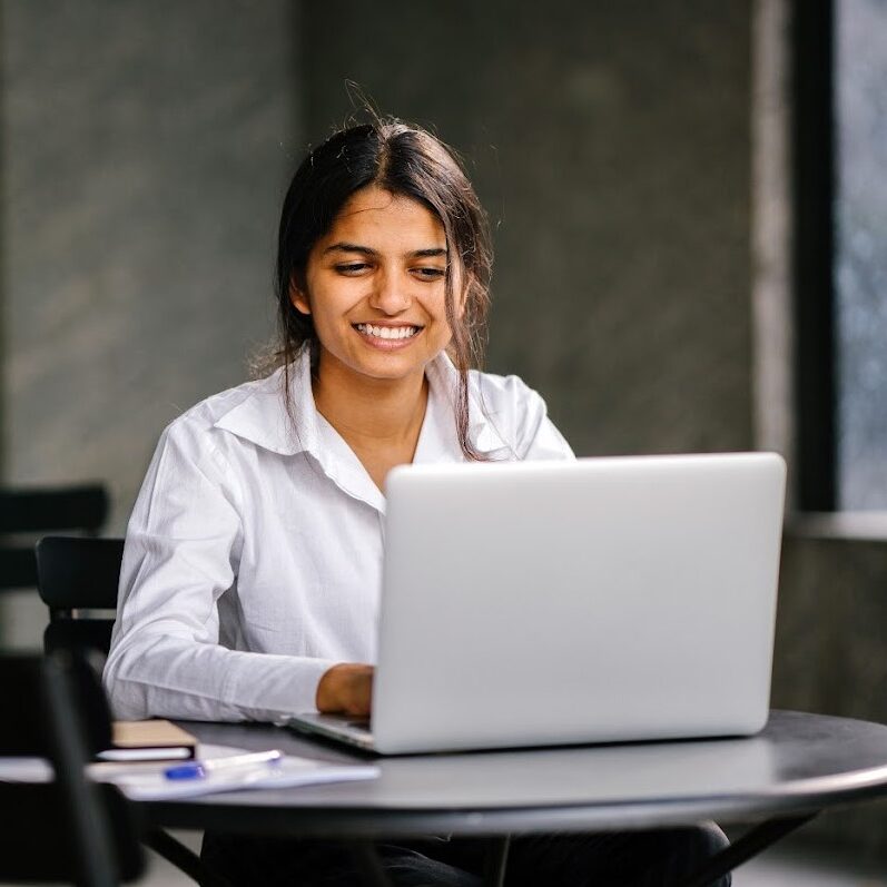 A young job-seeker applies for her job on her laptop computer. It is a portrait of her (Indian Asian woman) sitting at a table as she smiles and fills in her application.